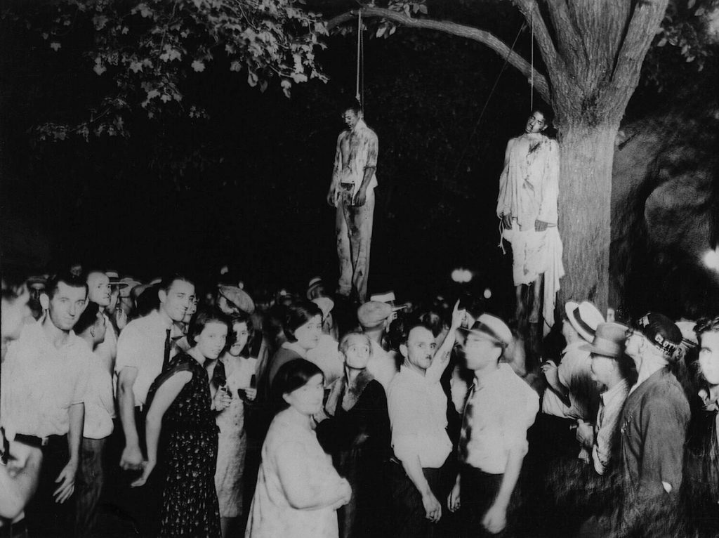 Historical black and white photograph depicting a group of people gathered under a tree where two individuals are hanging.